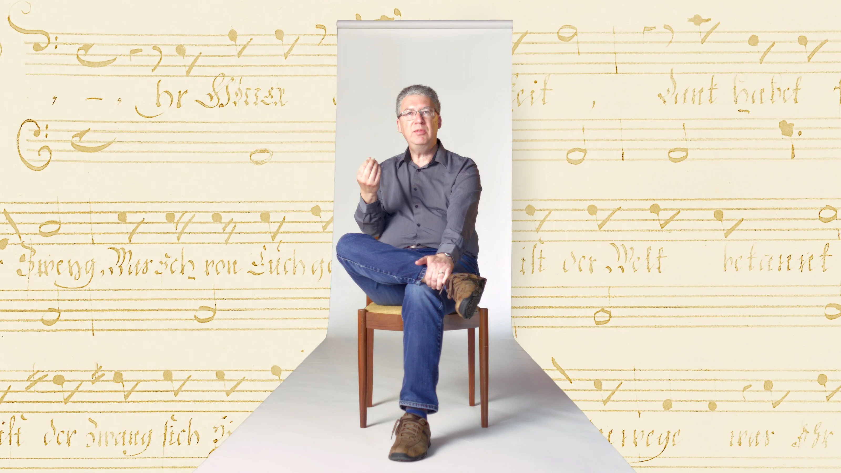 A man sits on a wooden chair with one leg crossed, gesturing with his hand, against a background of vintage sheet music.