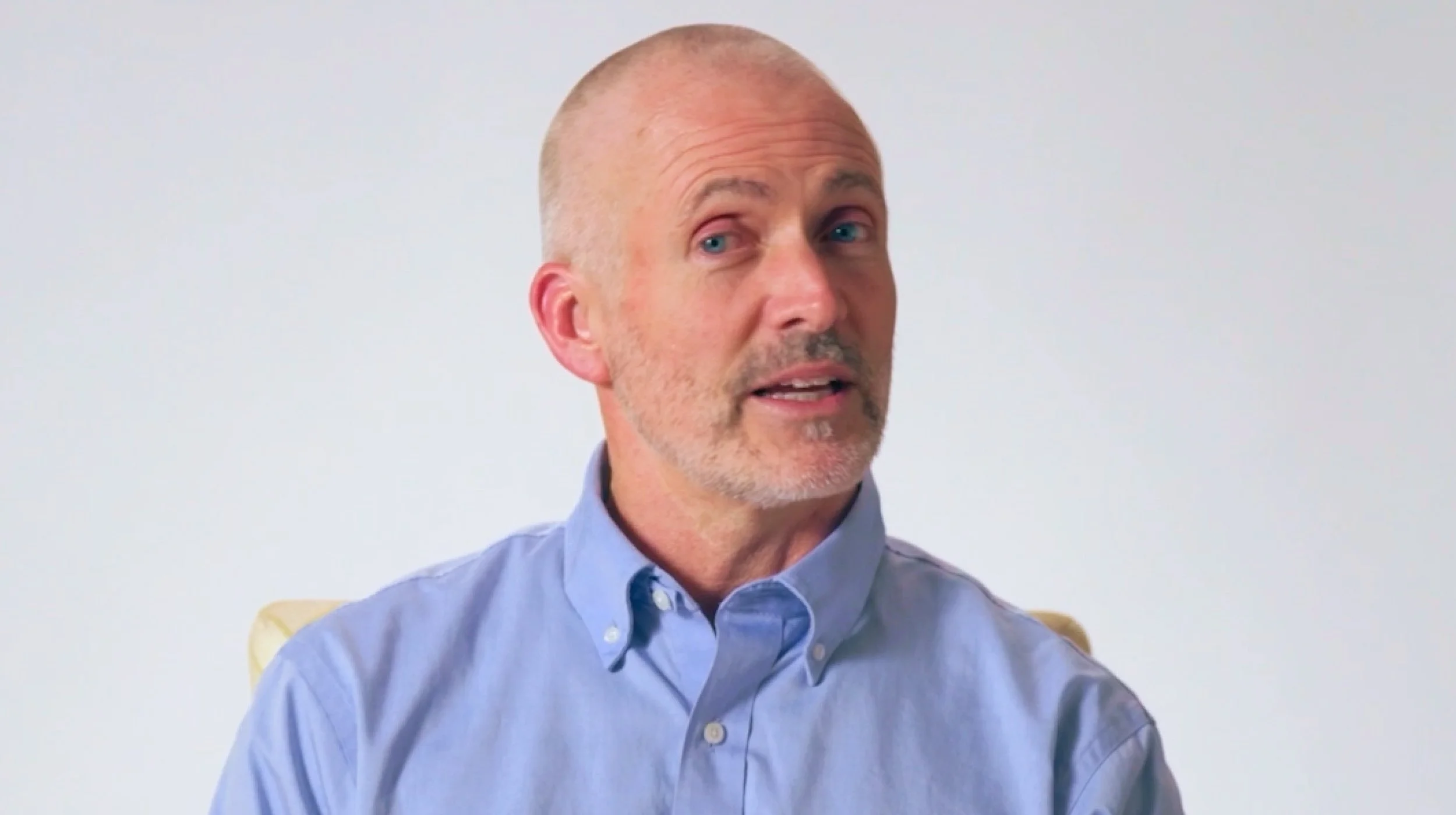 Bald man with a trimmed gray beard wearing a light blue button-up shirt, seated and looking slightly to the side against a plain light background.