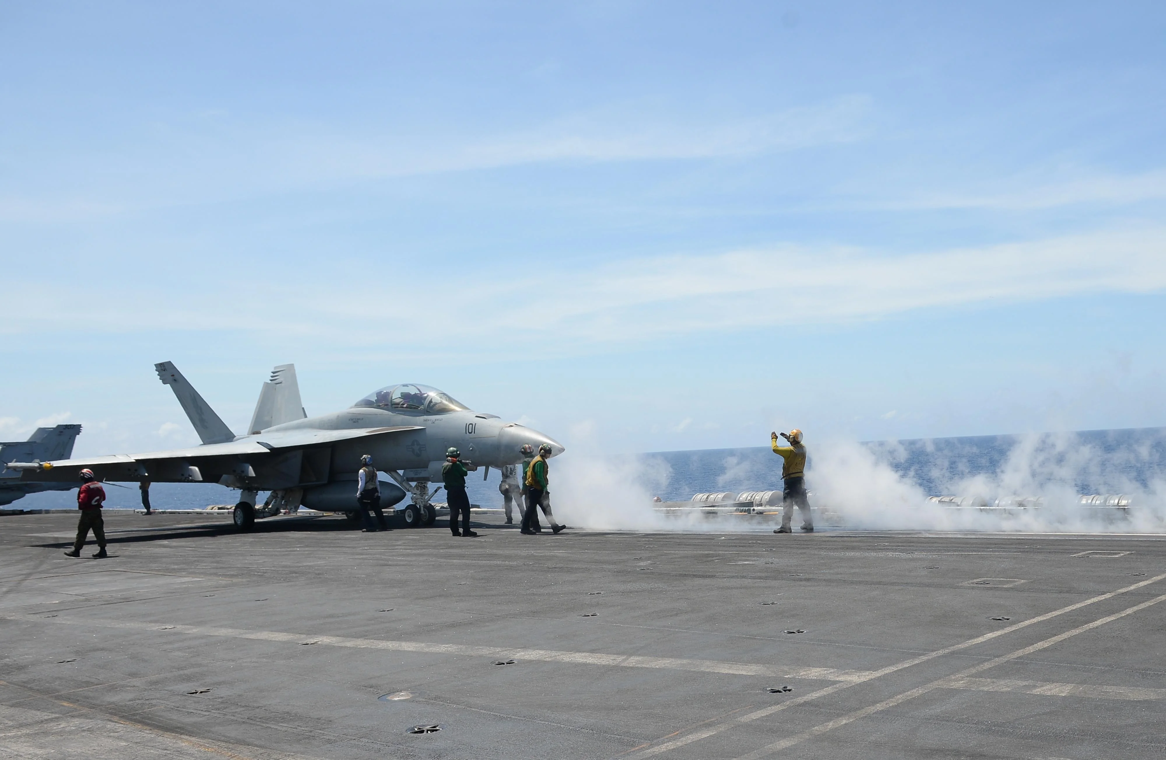 Sailors standing on the deck of a US aircraft carrier prepare to assist a jet for takeoff while steam billows around them.