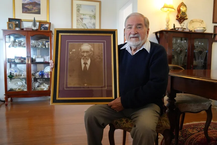 A man sits on a chair holding a portrait of his ancestor
