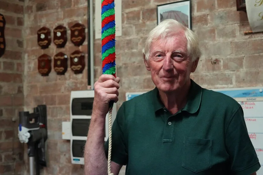 An elderly man stands and holds a rope that rings a bell.