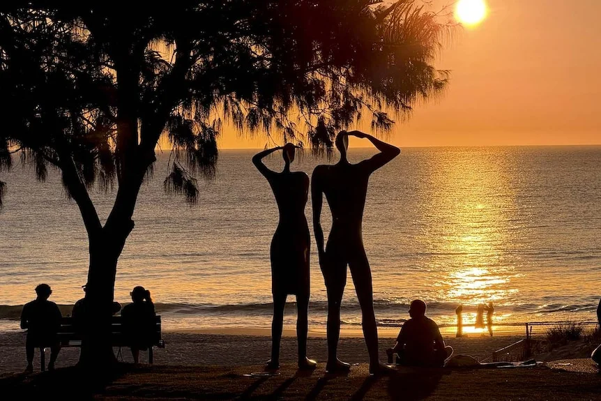 People sit at a beach among a statute as the sun turns the scene golden