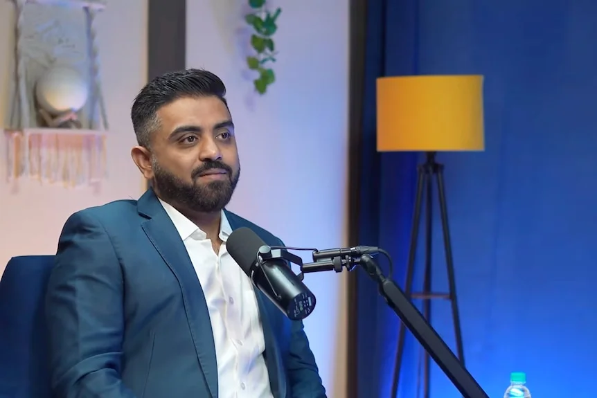 An Indian man sits in a chair and speaks into a vodcast microphone.