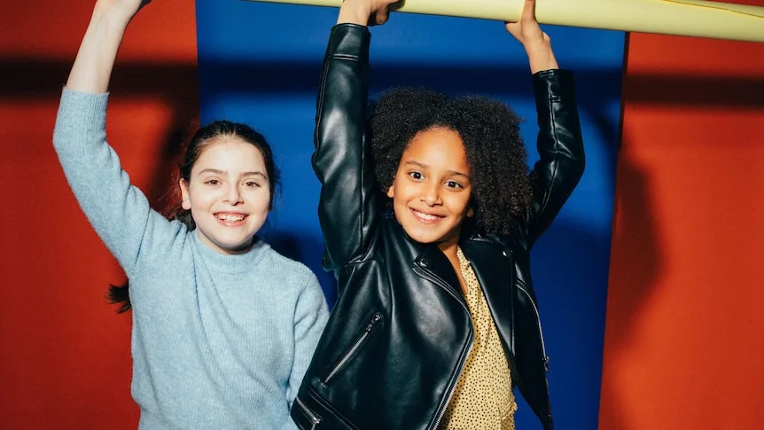 Two tween girls smiling with their arms raised holding on to playground equipment.