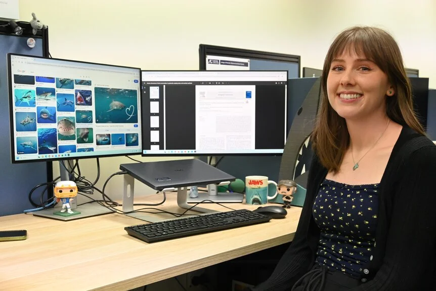 A woman sits at a desk with a computer screen showing pictures of sharks