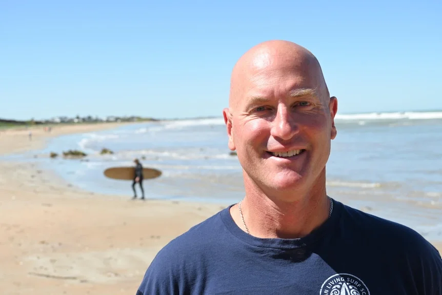 A man stands on a beach with a distant figure carrying a surfboard in the background