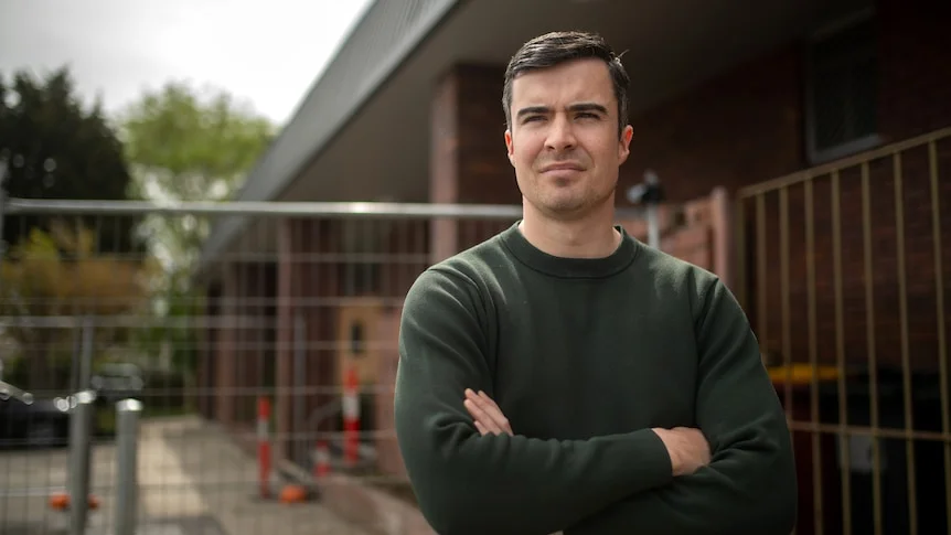Hayden stands in front of construction fencing with arms folded in a green jumper on a cloudy day.