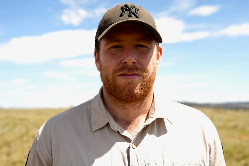 A man wearing a beige button-down shirt and black cap stands in a paddock smiling lightly.