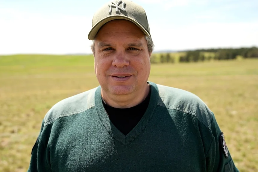 A man wearing a dark green sweater and beige cap stands in a paddock smiling lightly.