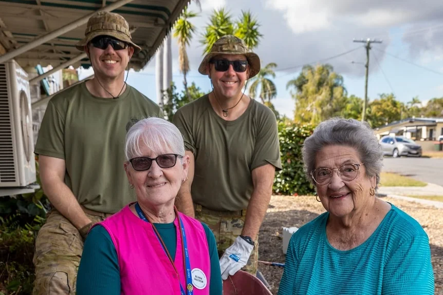 In a front garden, two older woman sit and smile at the camera in front of two standing soldiers in fatigues.