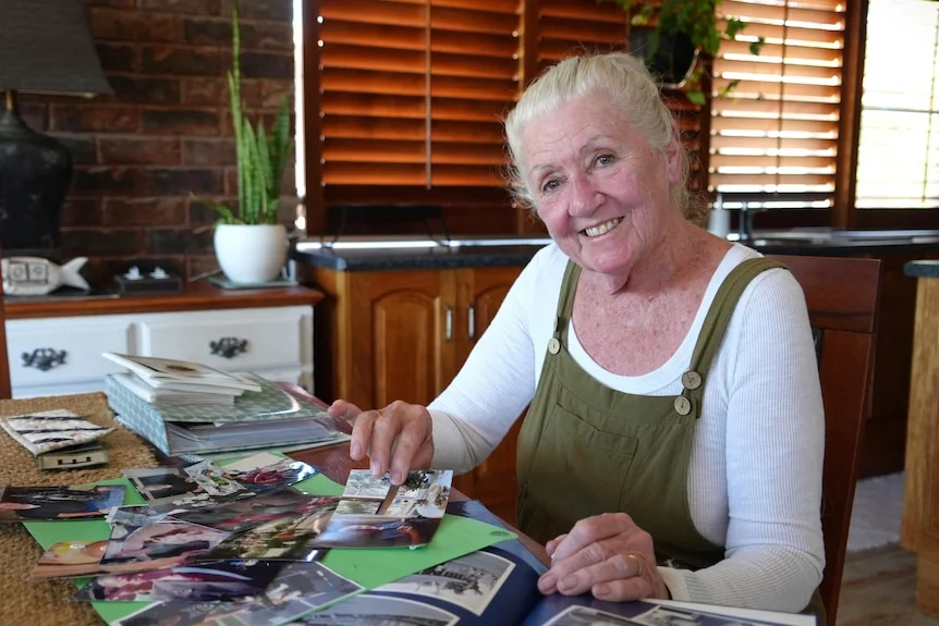 A mature woman wearing overalls sits at a table inside smiling at the camera, holding photos and clippings.