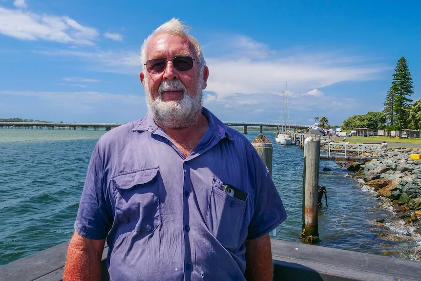 A grey-bearded man wearing a blue shirt and sunglasses stands in front of water.