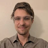 A headshot of a man with sandy brown hair and clear plastic glasses smiling at the camera. Blue collared shirt.