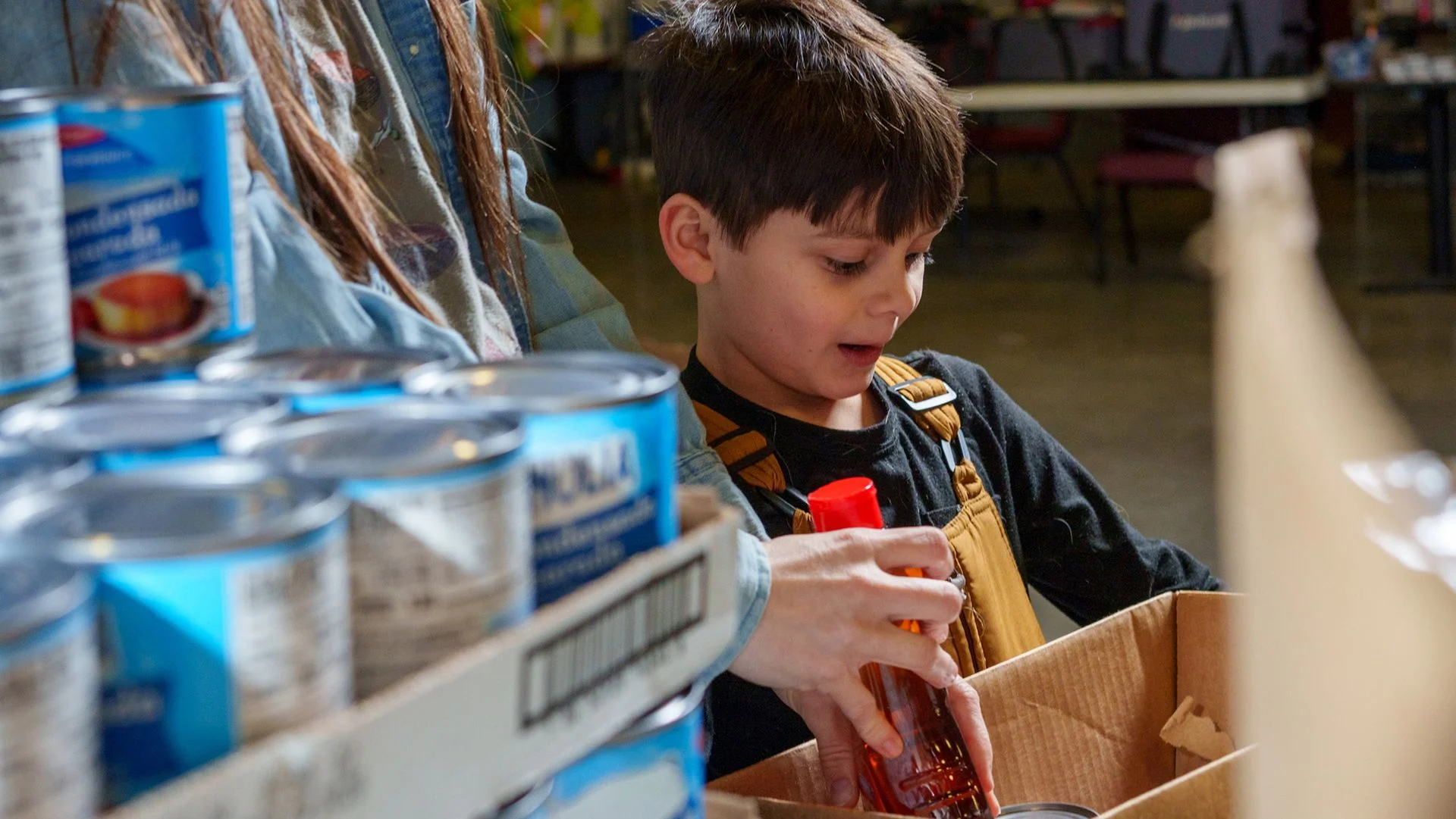 A boy holds a cardboard box with rows of cans in the foreground.
