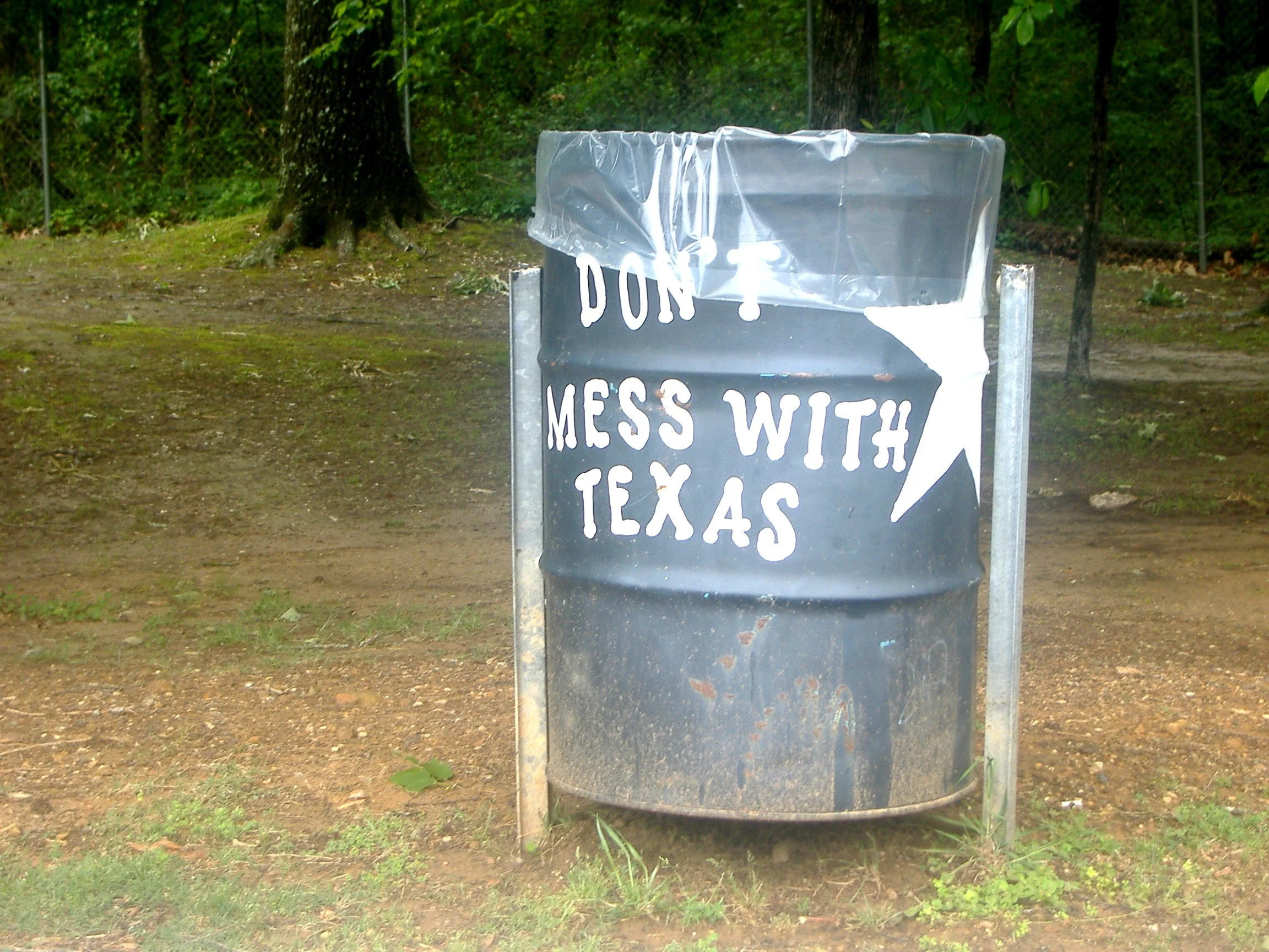 A black trash can in a wooded area with “DON’T MESS WITH TEXAS” and a white star painted on it.