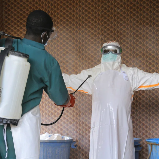 A health care worker in white protective clothing stands with their arms out while a colleague disinfects them inside a treatment center for Ebola patients in Congo.