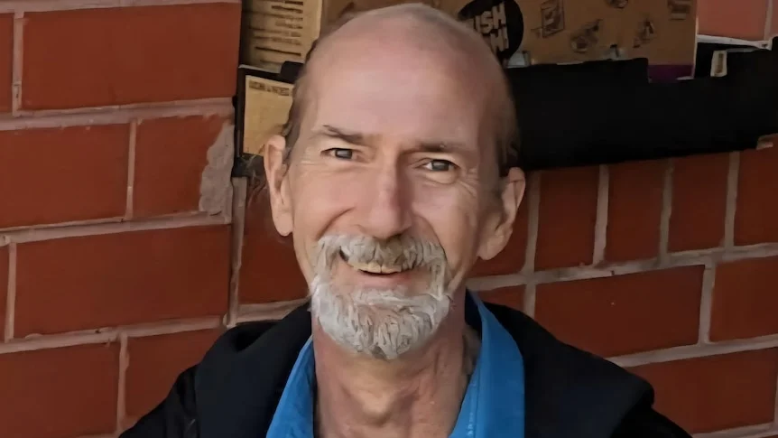 A balding Caucasian man with grey beard wearing a dark jacket and blue shirt