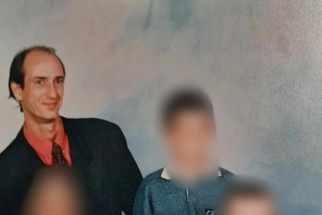 A man with receding hair wearing an orange shirt with dark suit and tie poses for a photo with others blurred