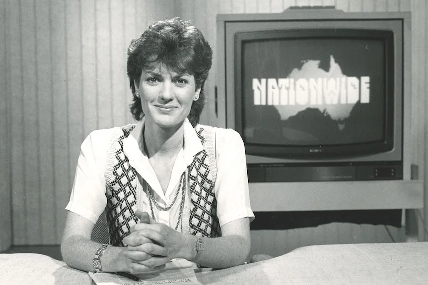 A black and white photo of a young woman sitting at a news presenter's desk with a TV behind her.