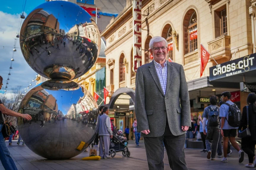 A man wears a grey suit, he has grey hair and stands in front of two polished silver balls in an outdoor shopping precinct