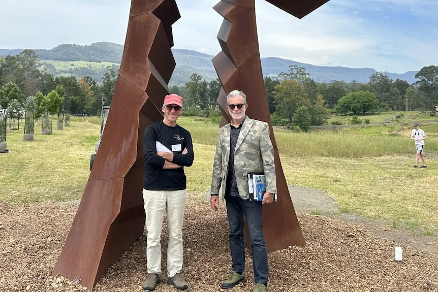 Photo of two men posing in front of a large steel sculpture in a field