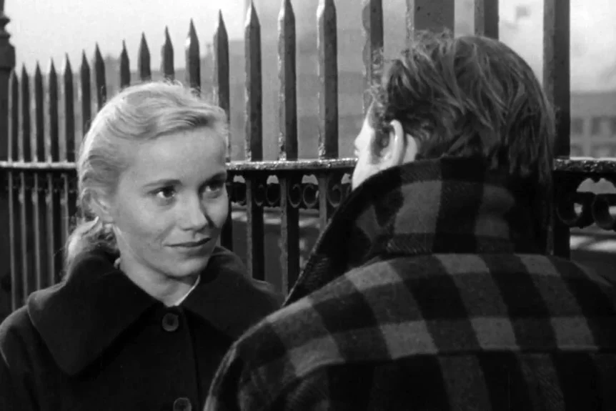 Black and white film still of Eva Marie Saint with her hair in a low ponytail, talking to a man in a thick jacket outside