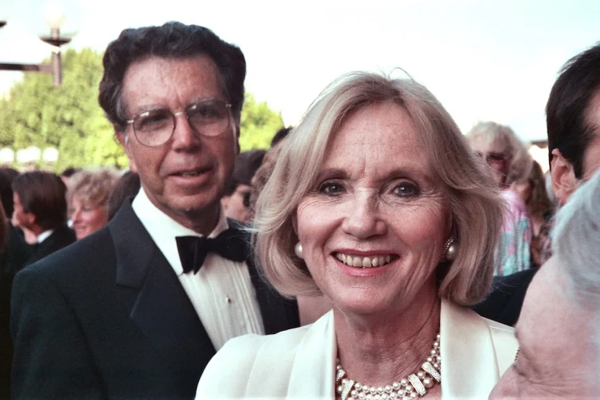 A middle aged Eva Marie Saint smiles at the camera wearing a pale suit and pearls, a man in glasses behind her