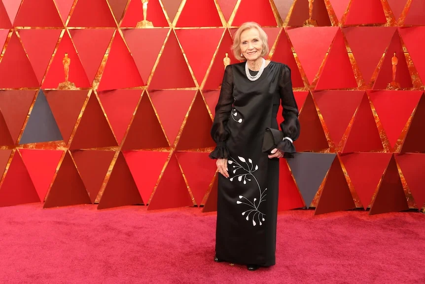 An older Eva Marie Saint in a long black satin dress and pearls, standing on the red carpet