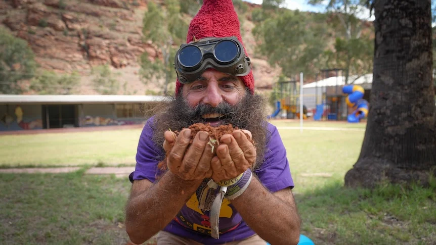 Costa Georgiadis is dressed as a garden gnome, wearing a pointed felt hat on his head. He has a big smile as his hands cup soil.