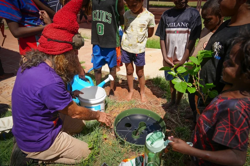 Costa Georgiadis kneels down, points to a piece of gardening equipment, a small group of young Indigenous kids huddle around.
