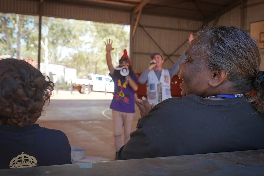Two children's performers addressing the crowd, an Indigenous woman can be seen smiling and clapping along in the foreground.