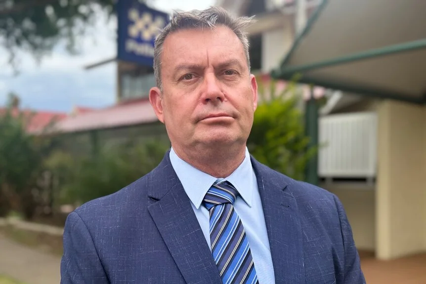 A man with a stern face stands in front of a police station.