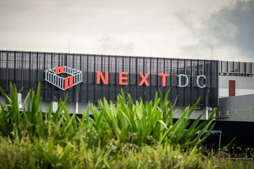 A sign on the side of a new building reads NEXTDC in grey and red font, greenery can be seen in a garden in the foreground.