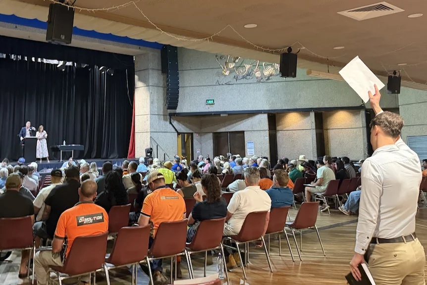A bidder raises a sheet of paper in a crowded Mount Isa auction room as an auctioneer stands on stage.