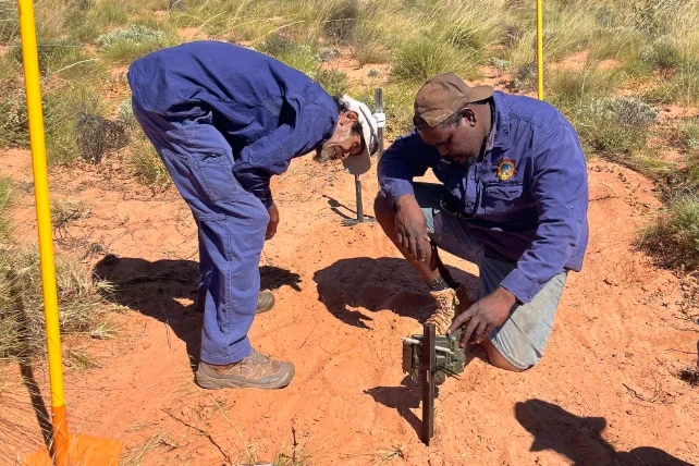 Two men dressed in dark blue ranger great setting up camera on red dirt.