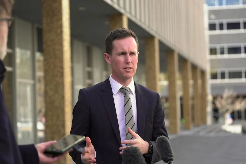 A man stands speaking into microphones outside, gesturing and wearing a suit and tie.