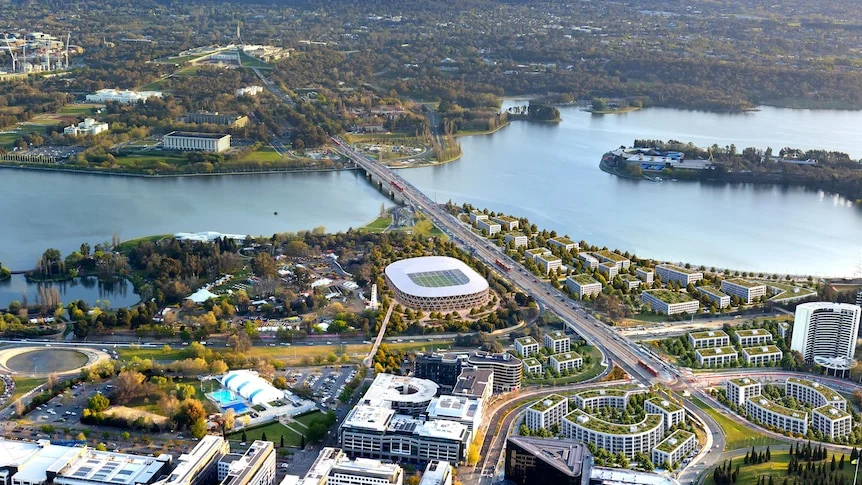 An artist render of a large stadium between a city centre and the edge of a lake with a double bridge across it.