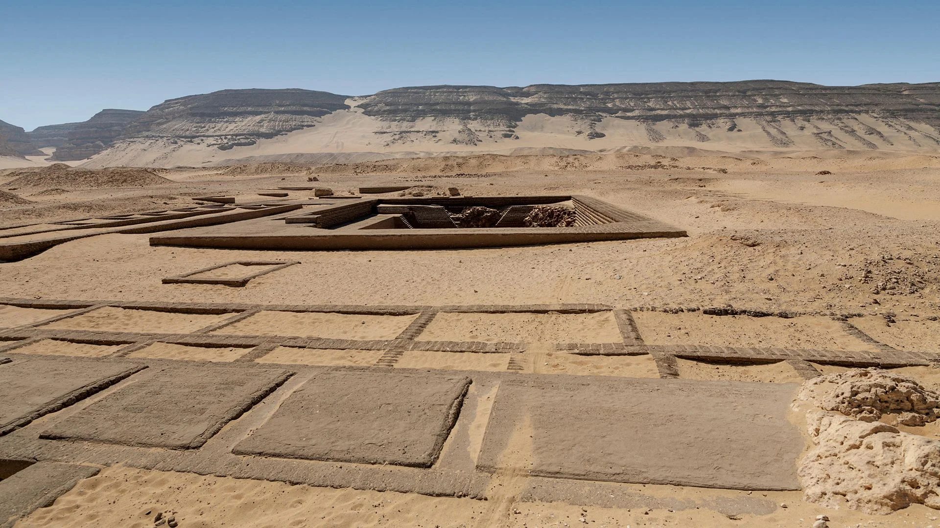 A series of square areas are seen in the desert, with a larger tomb in the background