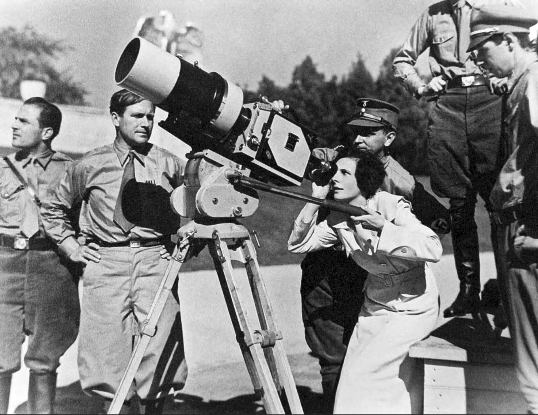 A woman looks through a large film camera on a tripod, surrounded by several men in military uniforms outdoors.