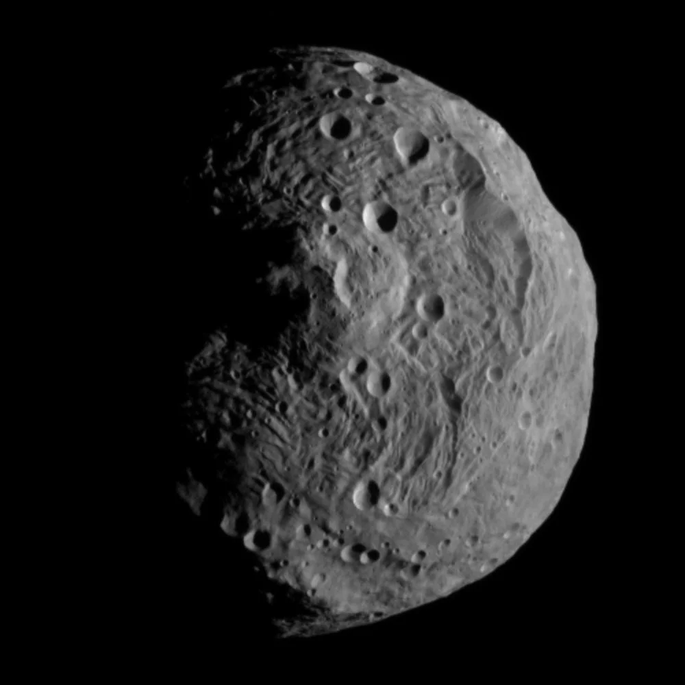 A gray, cratered asteroid or moon is shown against a black background, its surface covered in impact marks and ridges like those found near Olympus Mons, the highest mountain in the solar system.
