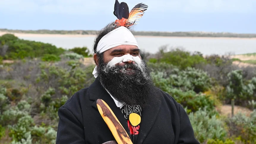 A man holding clapsticks, with face paint and feathers in his hair.