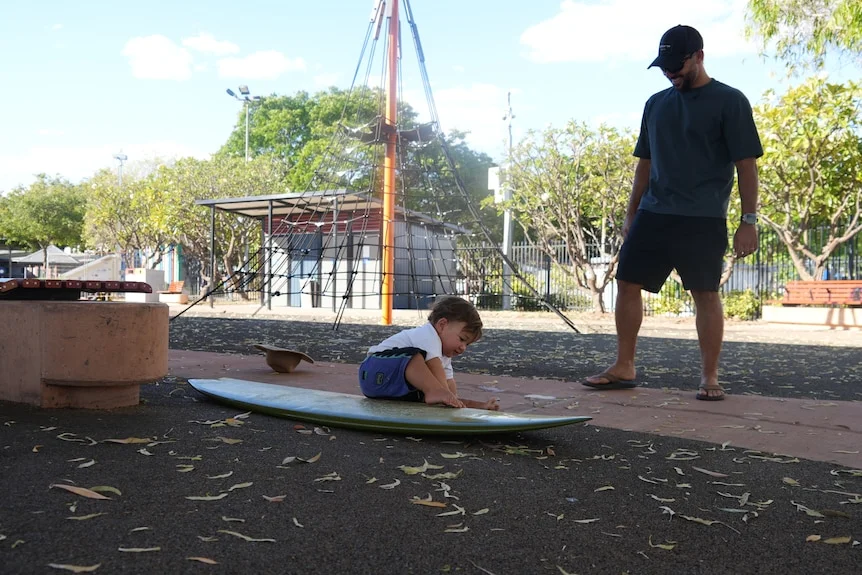 toddle sitting on surfboard with father watching behind, smiling