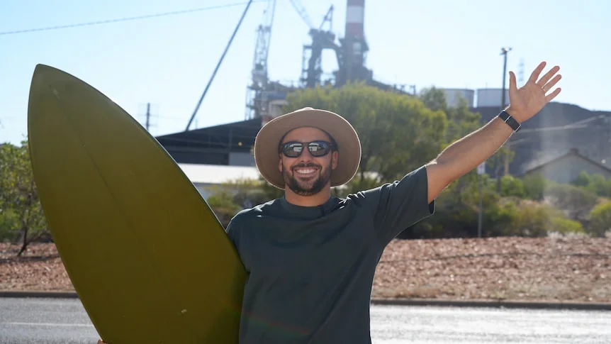 sufer stands holding surfboard wearing akubra hat in front of the mount isa mines