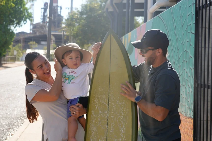 a wife, husband, and toddler standing in a suburban street smiling with a surboard between them