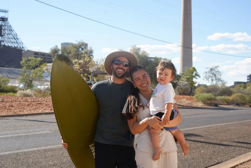 A man wearing a hat and sunglasses holds a surfboard as he stands alongside a woman carrying a child