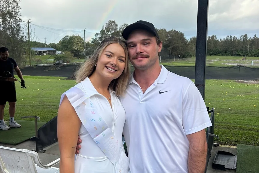 A man and woman wearing golf clothing standing at a driving range smiling at a camera