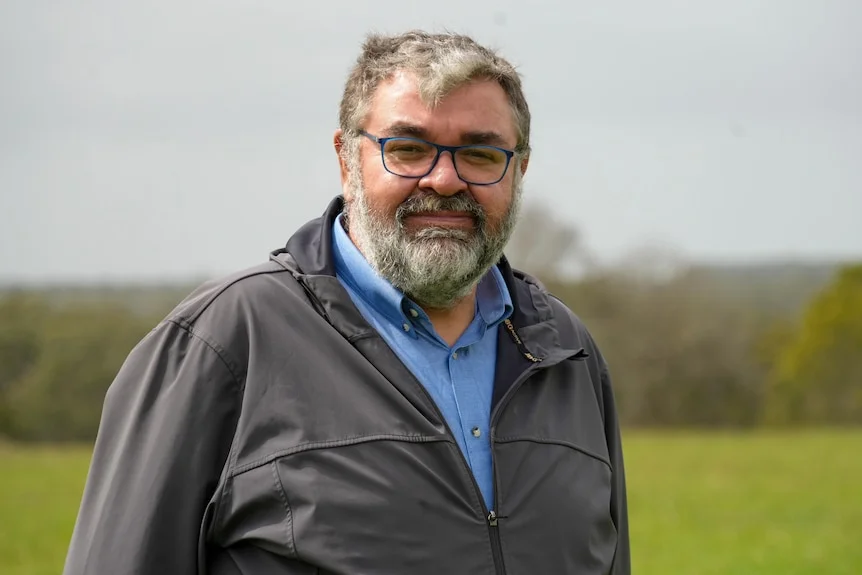 Damein Bell standing and smiling in front of the camera, a green landscape behind him.