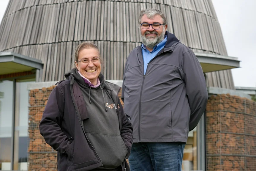 Evelyn Nicholson and Damein Bell stand smiling in front of a stone hut.