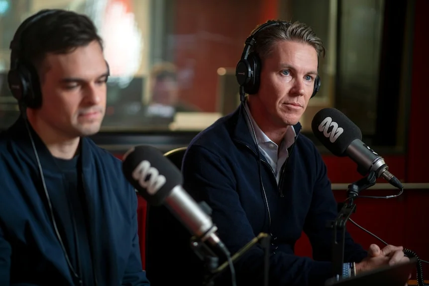 Two young men sit in a radio booth with red walls, wearing headphones and speaking into an ABC branded microphone.
