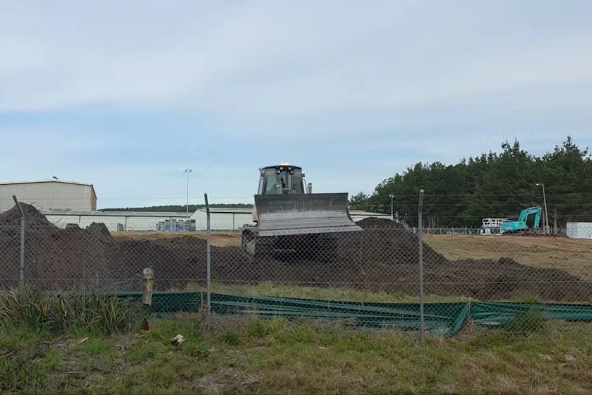 An excavator on a pile of dirt, behind a fence.